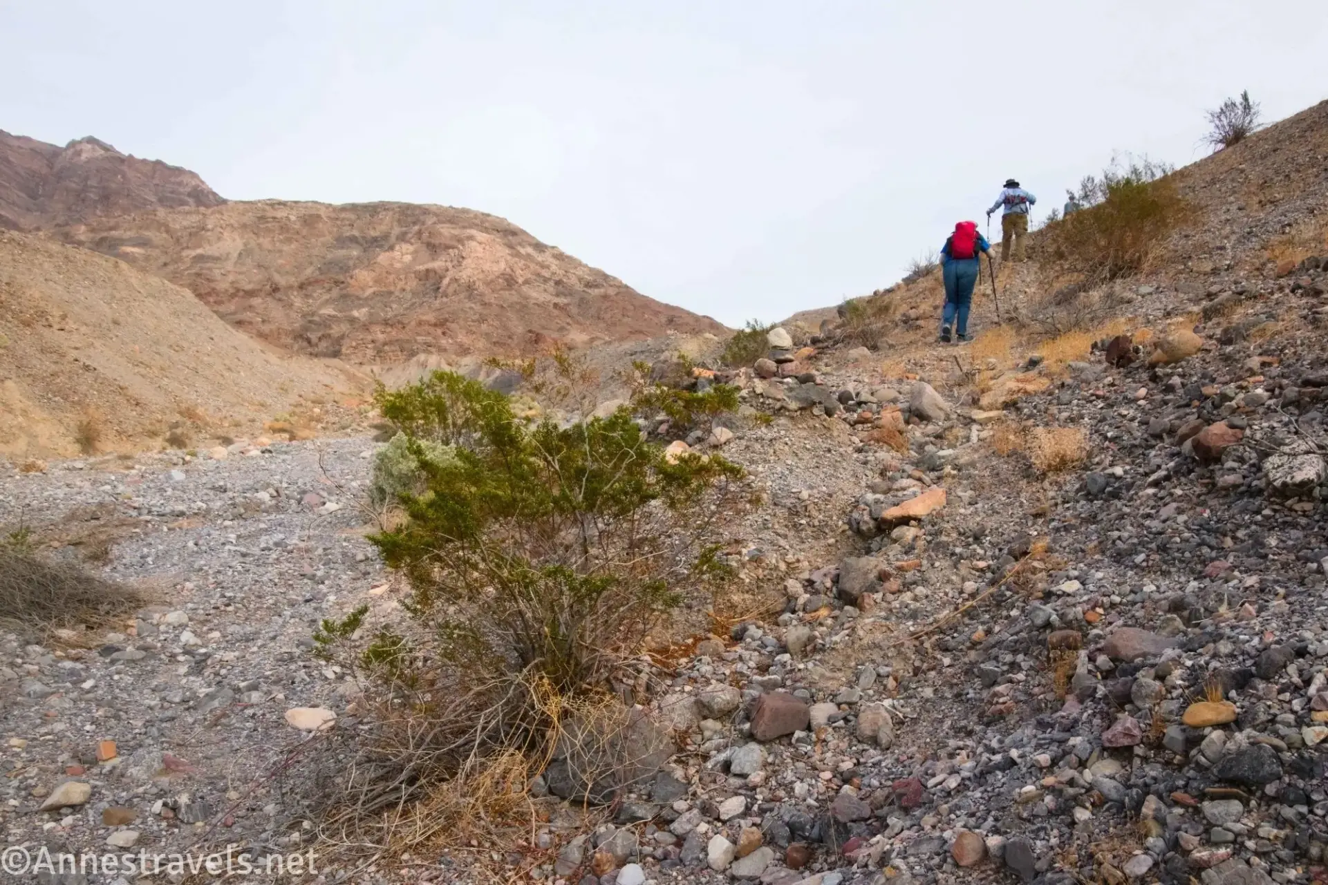 Trail into the Wash A gravel hillside with desert plants and two hikers beside desert hills