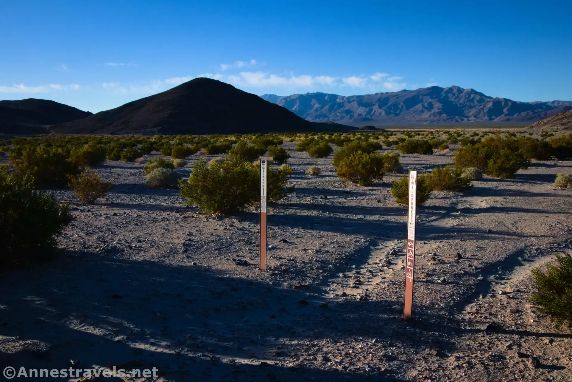 Beginning of the Lake Hill Hike Two vertical signs bar vehicles from a brushy desert plain with a dark hill and distant mountains