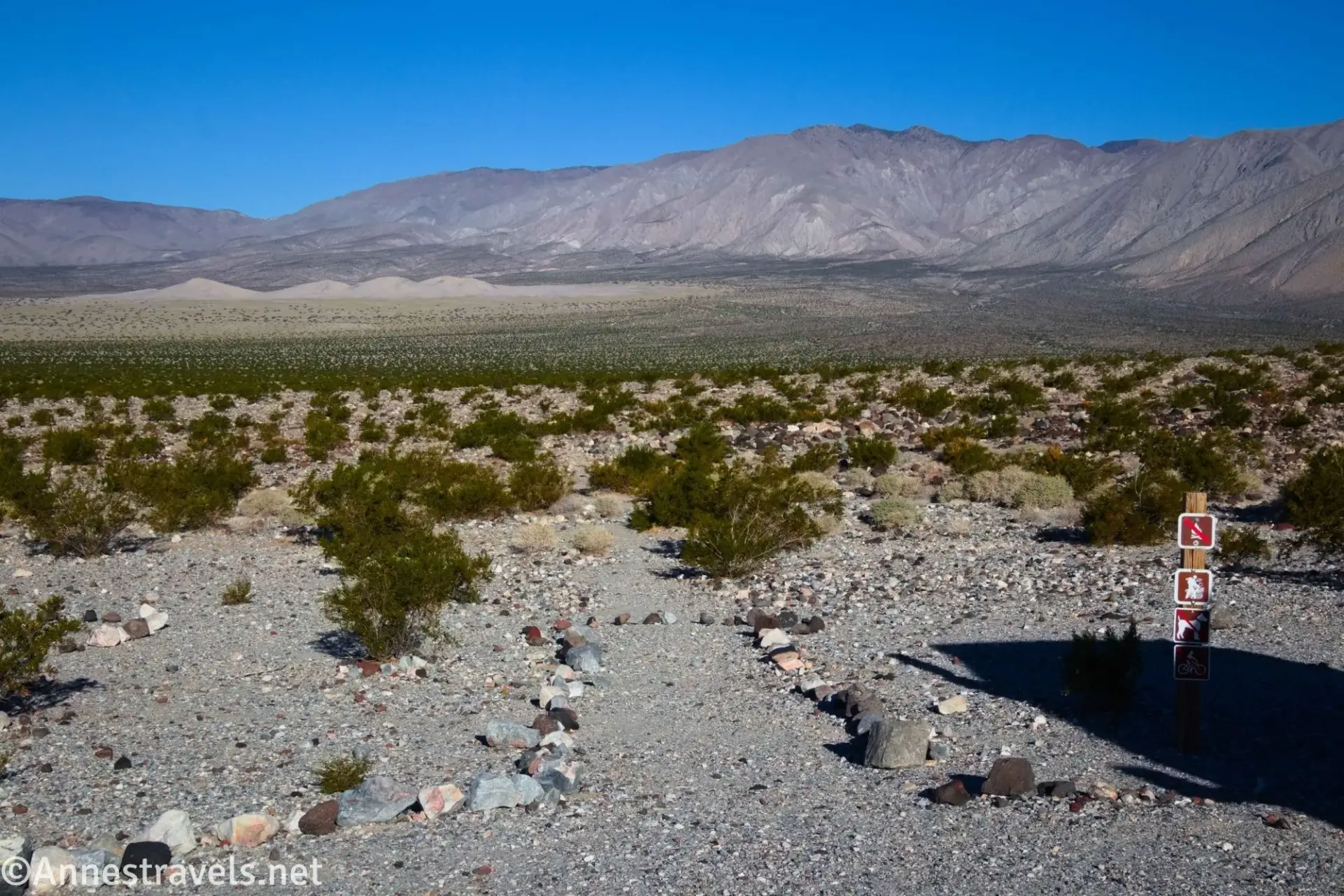 Trailhead for Panamint Dunes A sign beside a row of rocks on a brushy plain with sand dunes and mountains in the distance