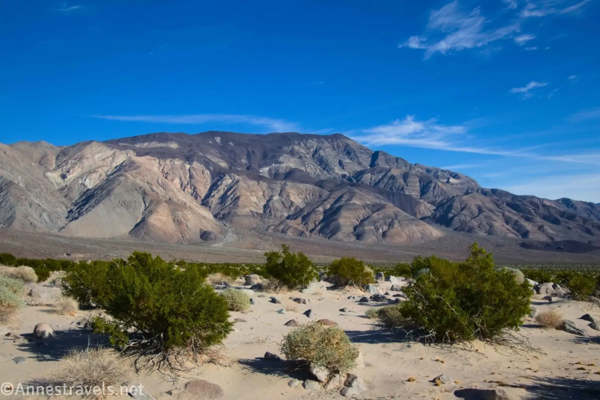 Cottonwood Mountains Brush on a desert plain with striped desert mountains below a few clouds in a blue sky