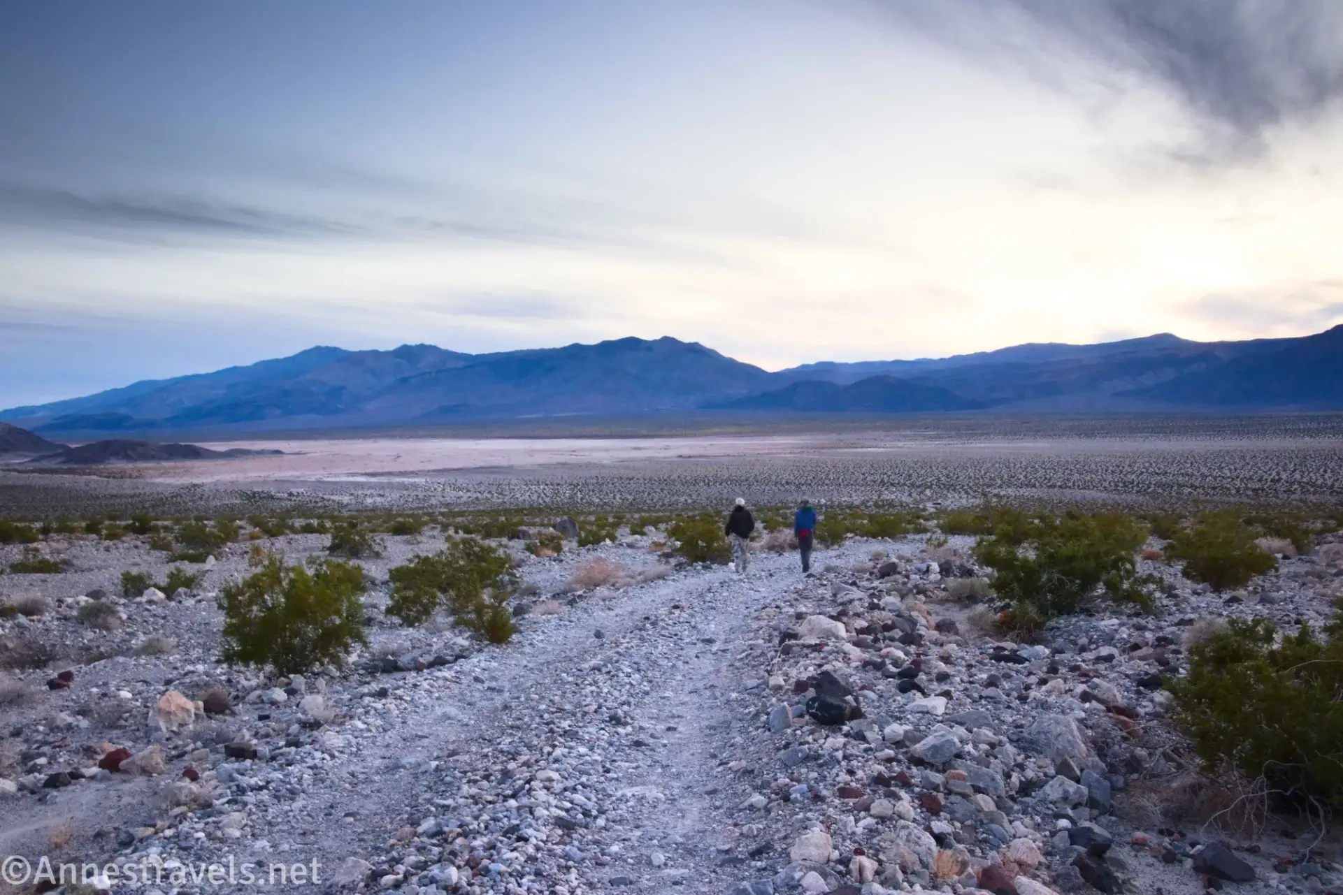 Two hikers on a dirt and gravel road above a desert plain and below dark clouds