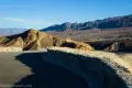 Stone wall along the back side of the paved Zabriskie Point viewpoint with a yellow hill and distant gray desert mountains in Death Valley National Park
