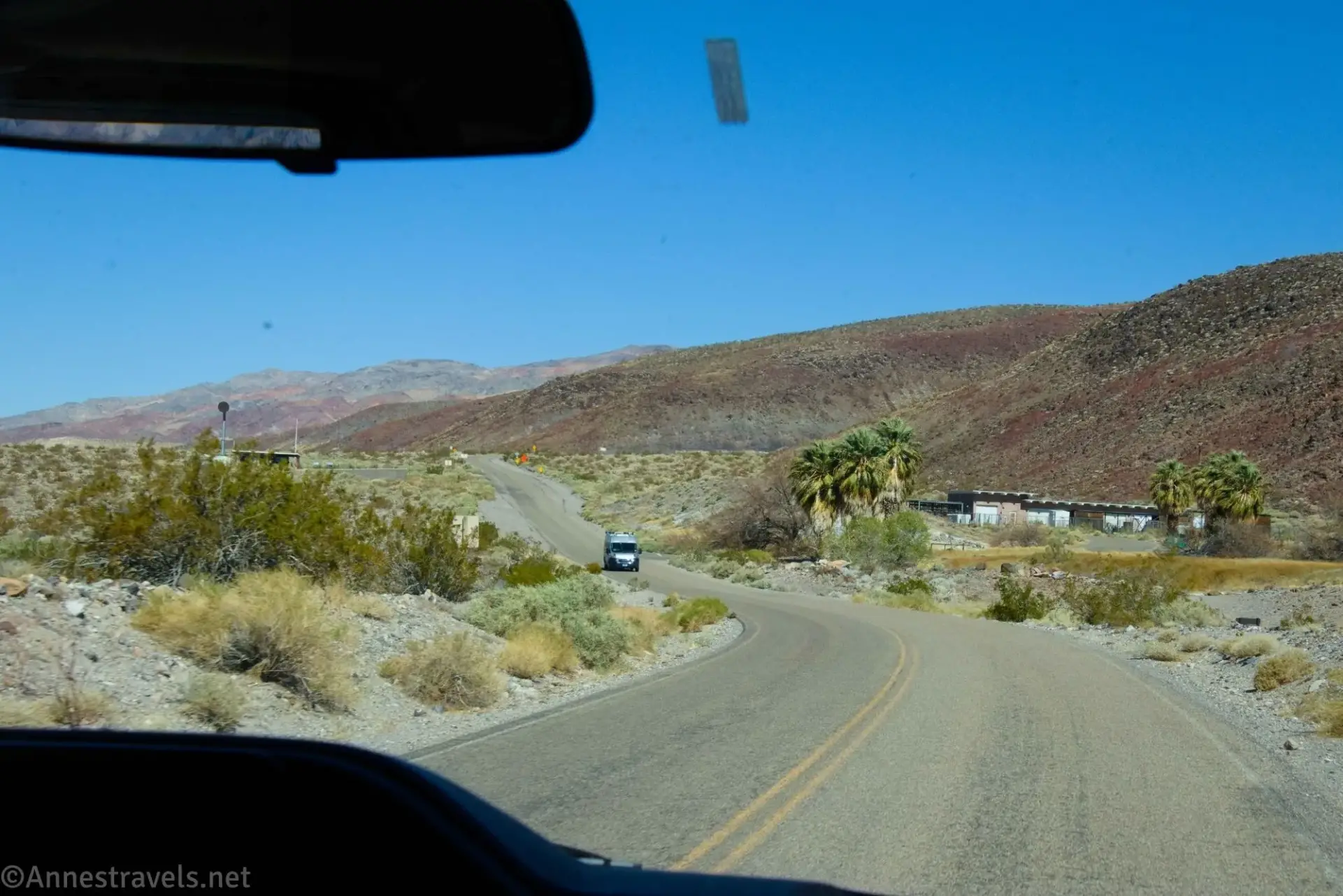 The Grapevine Ranger Station and palm trees on either side of a dusty paved road with a double yellow line and an oncoming camper van