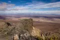 A view of a rocky outcrop above a desert valley with a few green fields, mountains, and clouds in a blue sky