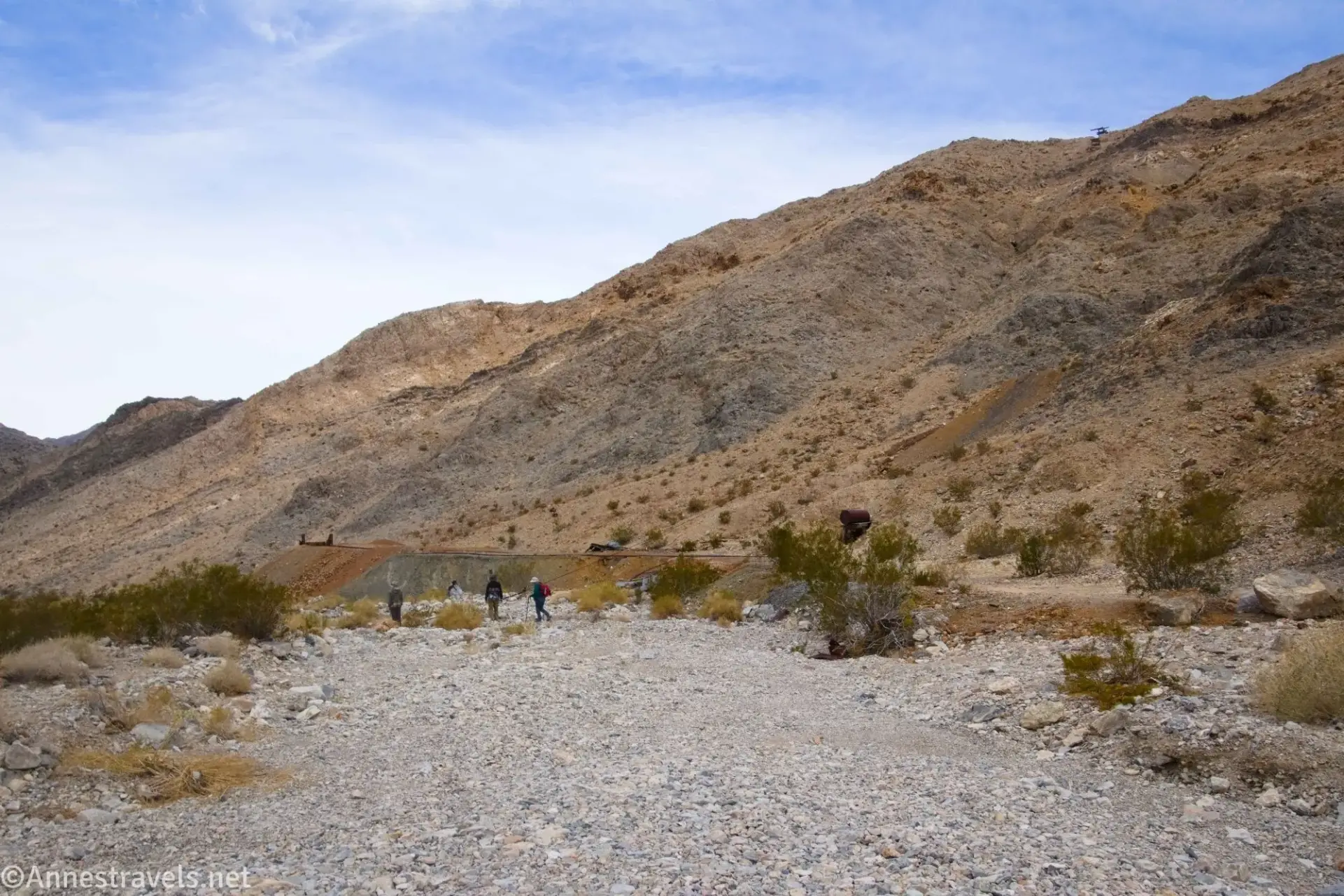 Brown mountains above a brown and gray slag pile and a gravel wash with three hikers