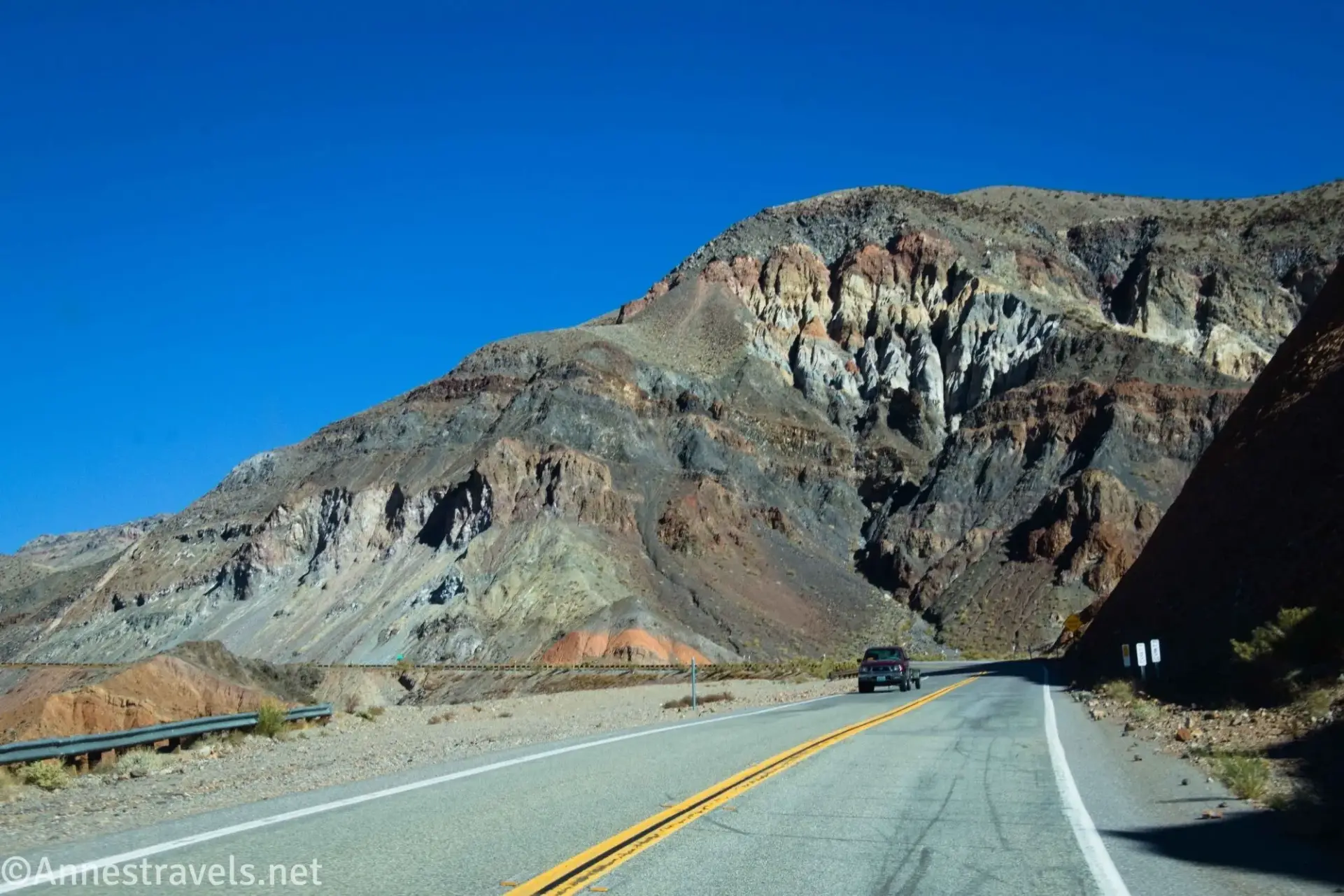 A vehicle on a paved road below a colorful badland hill
