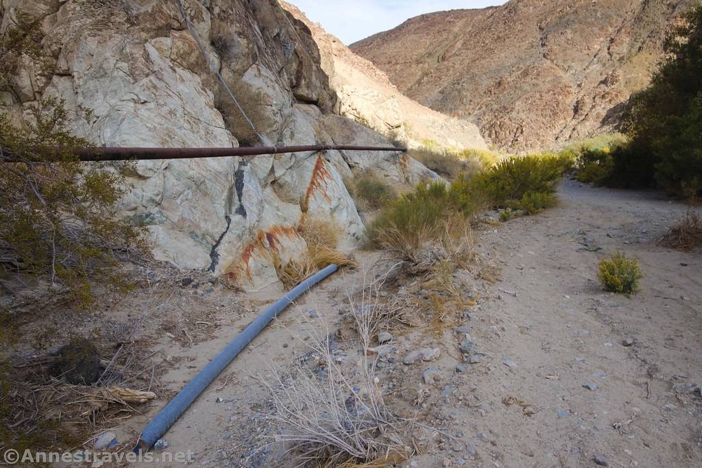 A pipe on the canyon wall and another pipe on teh ground beside a dusty trail in a desert canyon