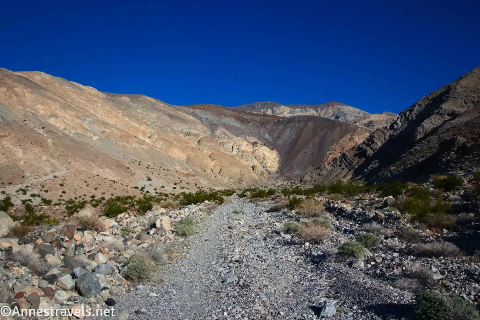 A gravel road between bushes and rocks heads for colorful cliffs of a canyon