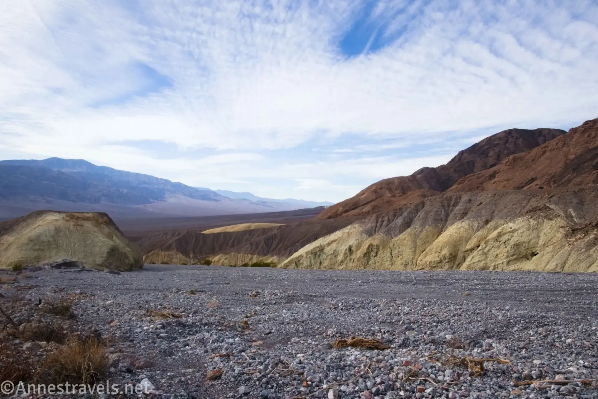 Clouds over red, brown, and yellow badlands and a gravel wash