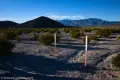 Beginning of the Lake Hill Hike Two vertical signs bar vehicles from a brushy desert plain with a dark hill and distant mountains