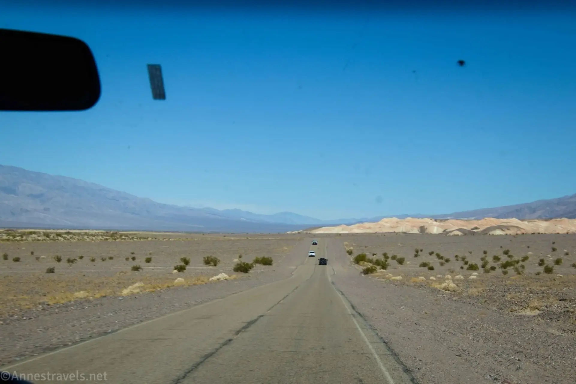 A dusty paved road stretches across the desert with three cars on it toward tan hills