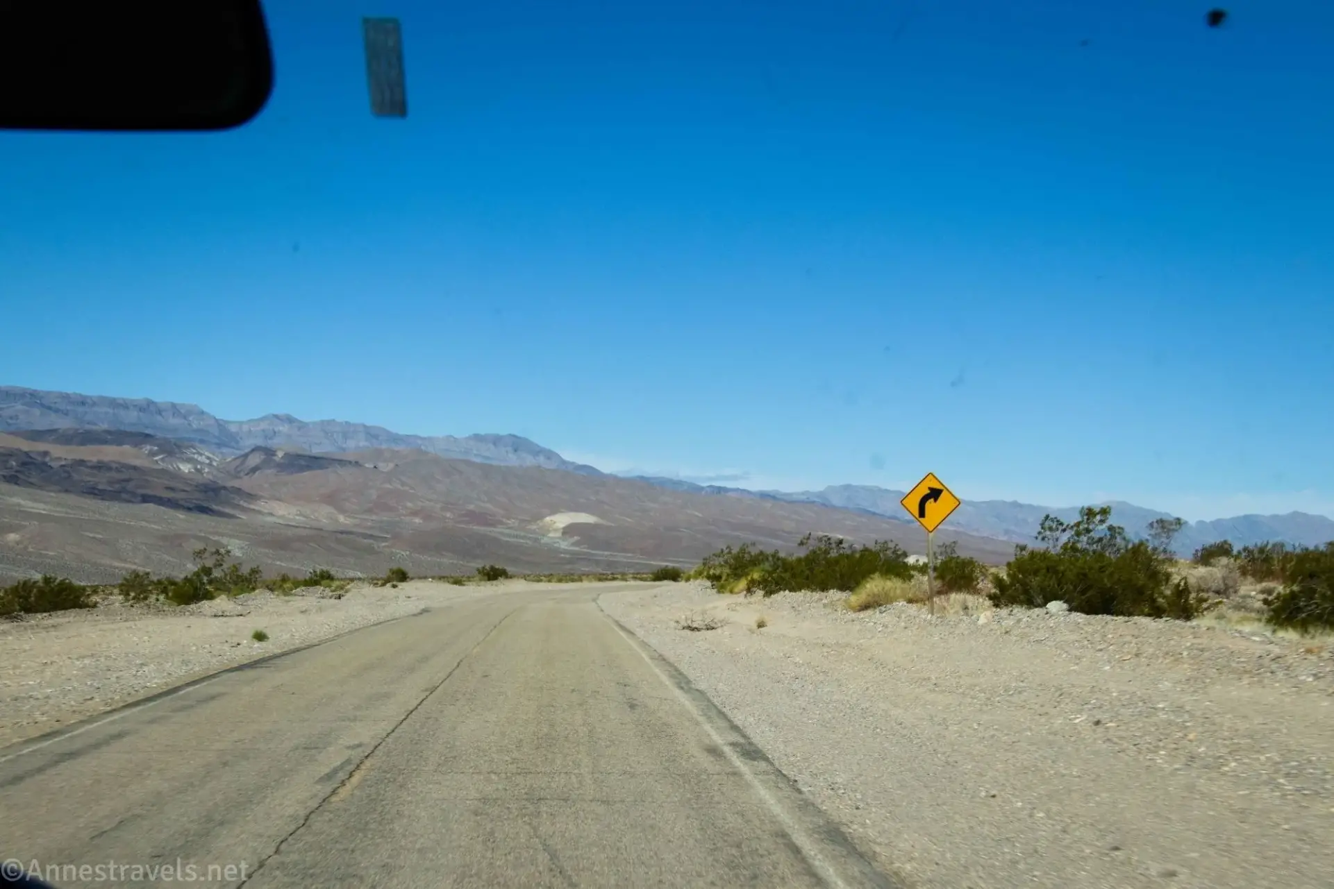A dusty paved road between gravel shoulders with a yellow curve sign to the right of the road