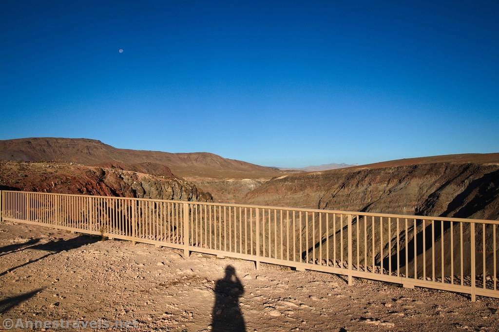 A fence along the edge of a canyon and shadow on the ground with the moon in the sky