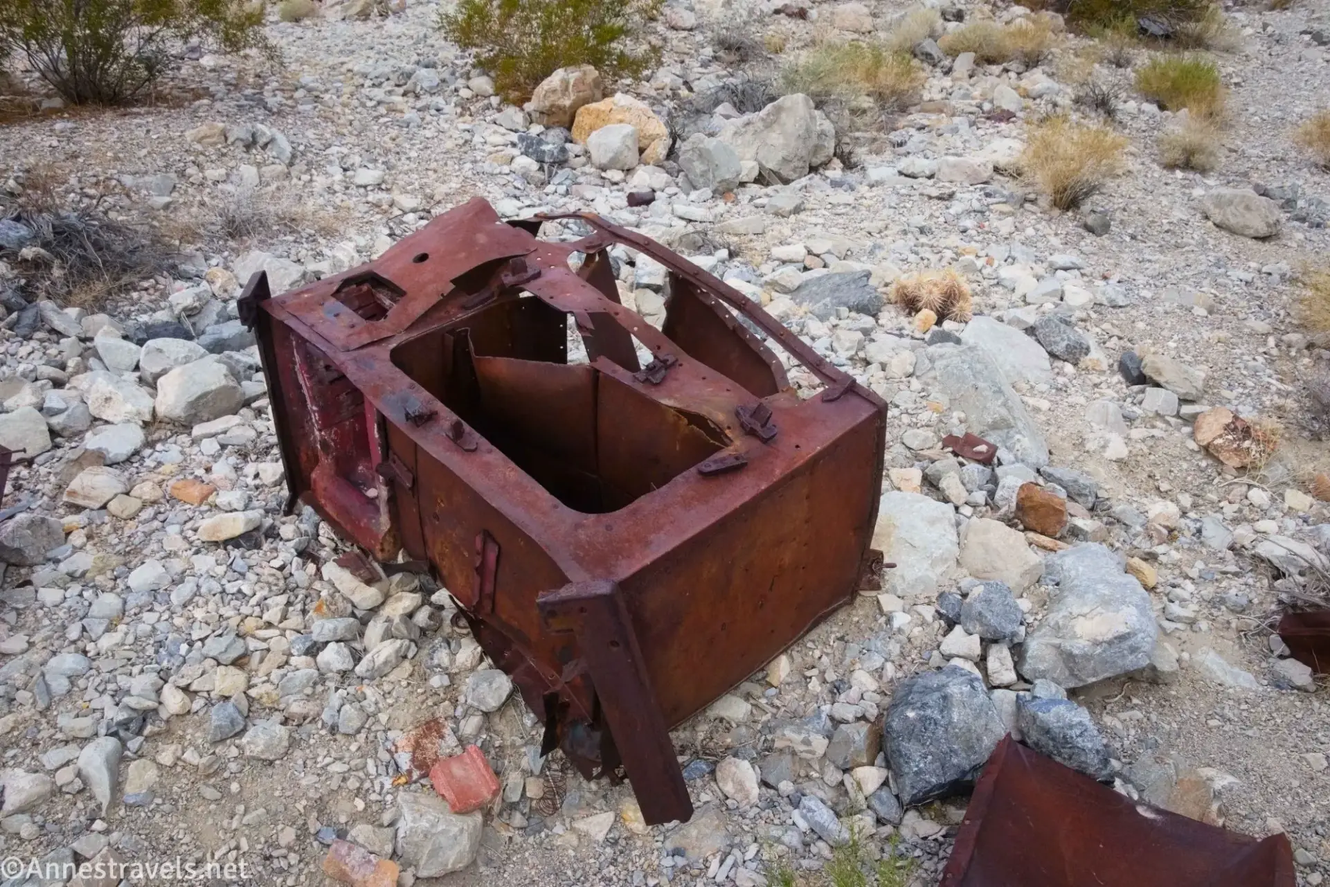 The rusting remains of a stove on a gravel surface with desert brush