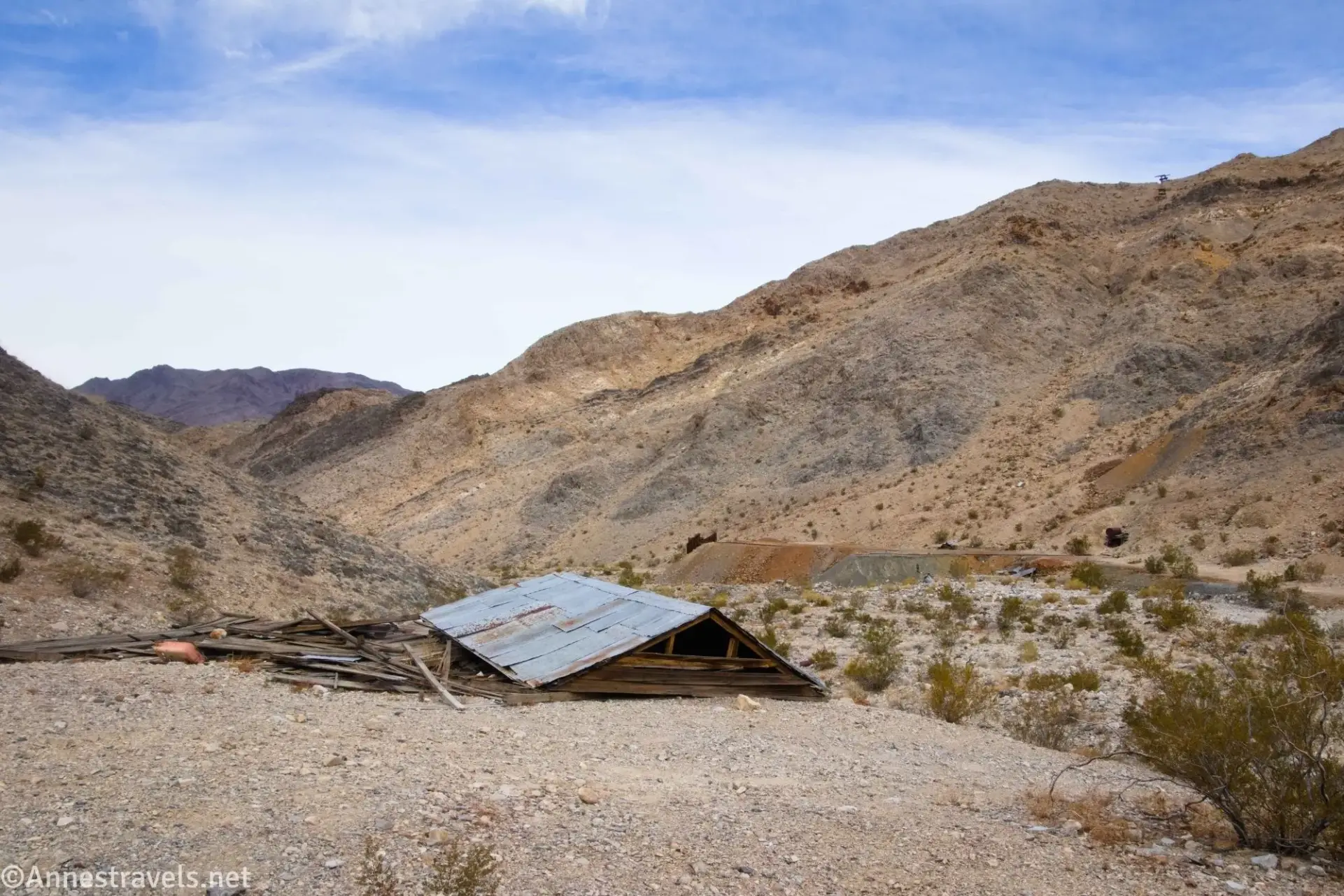 A collapsed building on a gravel area with a brown-streaked talus pile below brown mountains