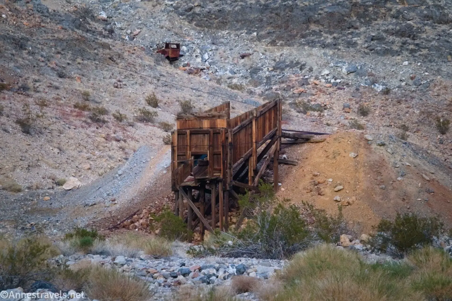 A brown wooden ore bin beside a lighter brown slag pile below a rocky hillside with a truck perched on the hill