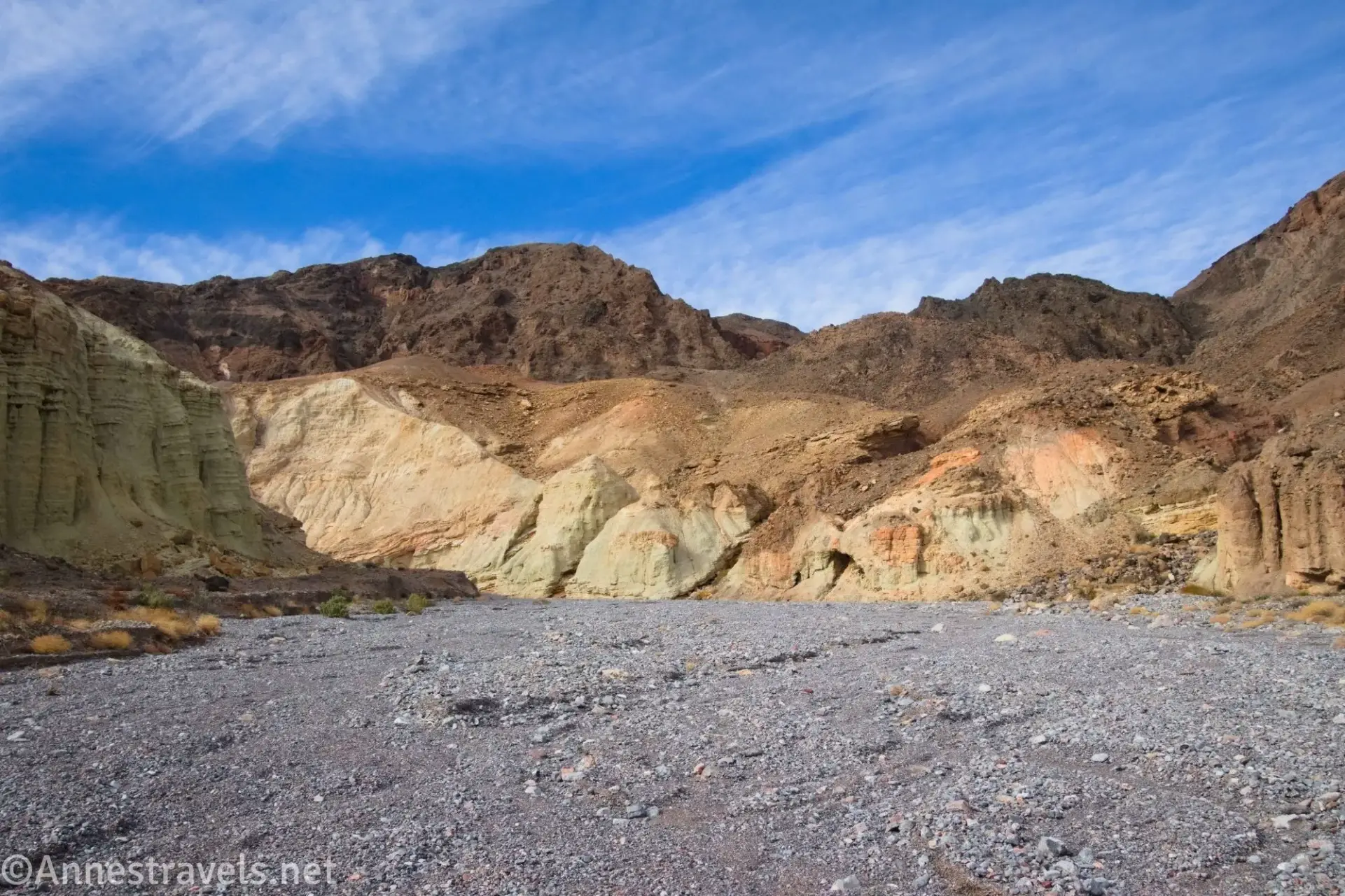 Yellow badland canyon walls with brown mountains behind and a gravel streambed in the foreground